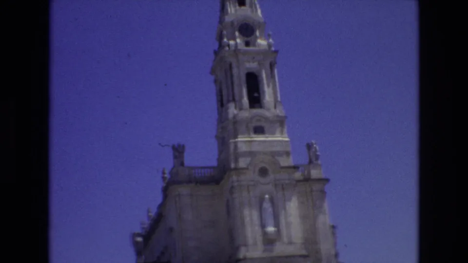 1972 Portugal 8mm frame with a low-angle view of a church tower, arched openings, roofline statues, and pale stonework under a saturated blue sky.