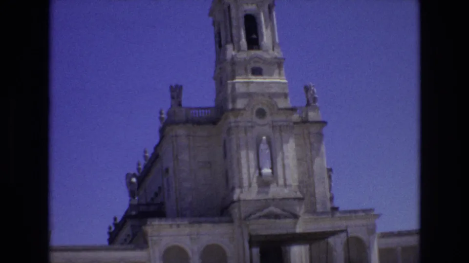 1972 Portugal 8mm frame showing the church tower and facade from below, with arched windows, stone ornament, a statue niche, and visible dark borders from the scan.