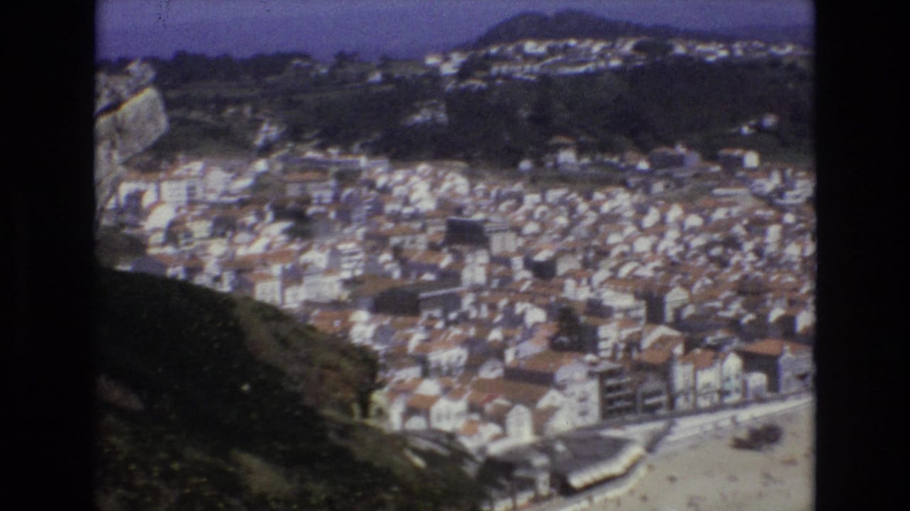 1972 Portugal 8mm frame looking down from a high vantage point over a seaside town, with red-tiled roofs, beach, surrounding hills, and a cross visible on the left ridge.