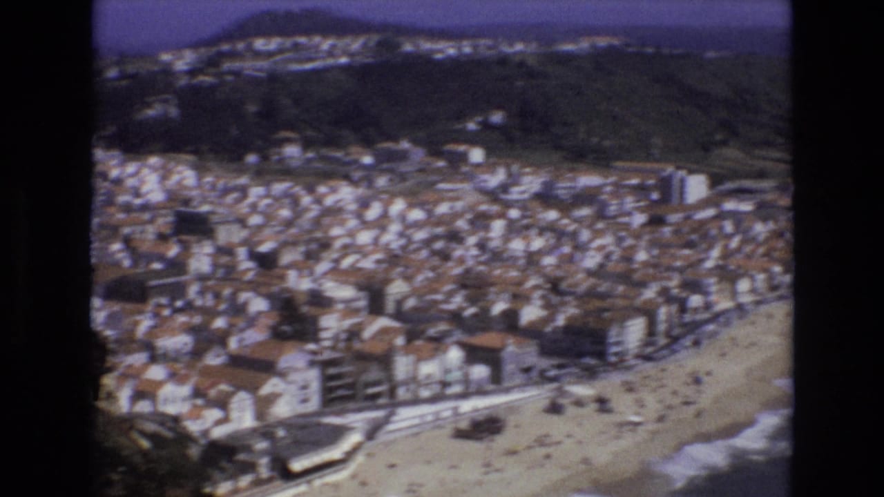 Coastal town seen from above with beach, waterfront buildings, hills, and ocean.