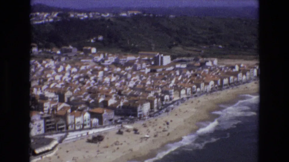 1972 Portugal 8mm frame of a beachfront town below green hills, with tiny beachgoers on the sand, a row of buildings along the shore, and waves breaking at the right.