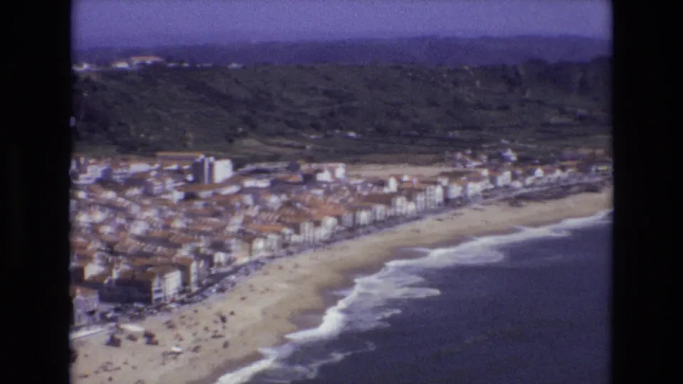 1972 Portugal frame from an 8mm home movie showing a wider sweep of beach, ocean waves, waterfront town buildings, green hills, and hazy mountains beyond.