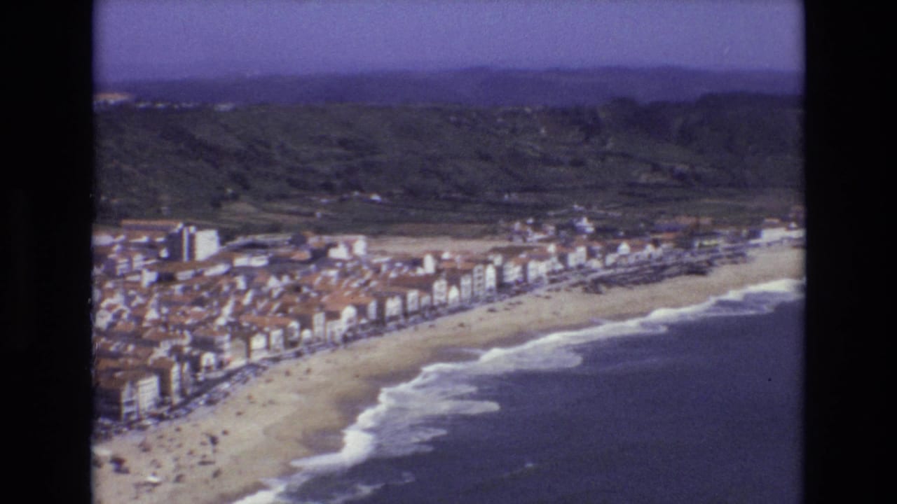 1972 Portugal 8mm aerial view of a seaside town lining a curved sandy beach, with surf, green hills, and a hazy sky visible beyond.