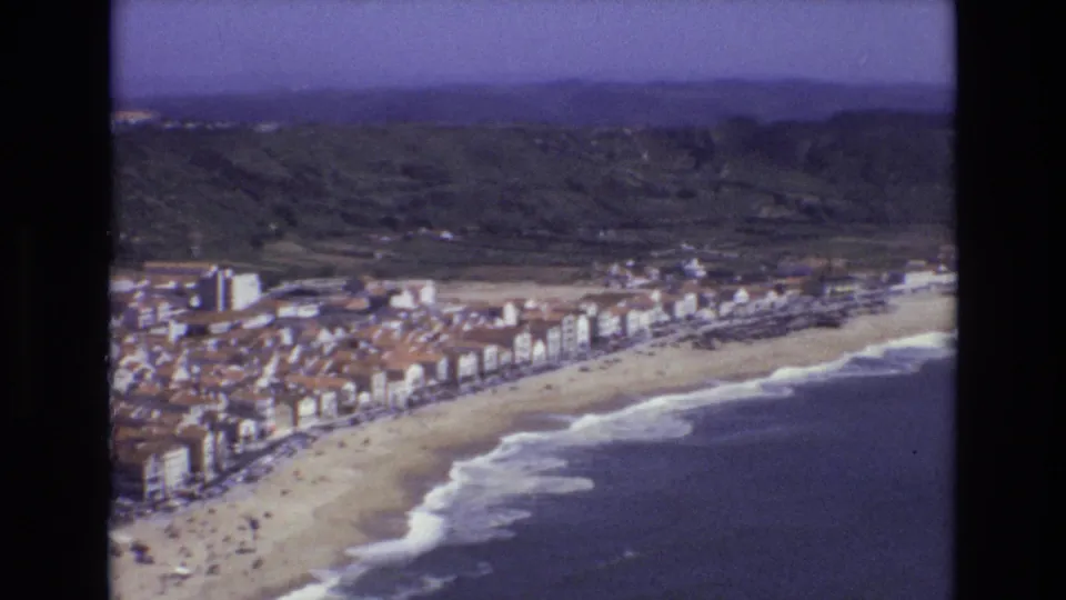 1972 Portugal 8mm aerial view of a seaside town lining a curved sandy beach, with surf, green hills, and a hazy sky visible beyond.