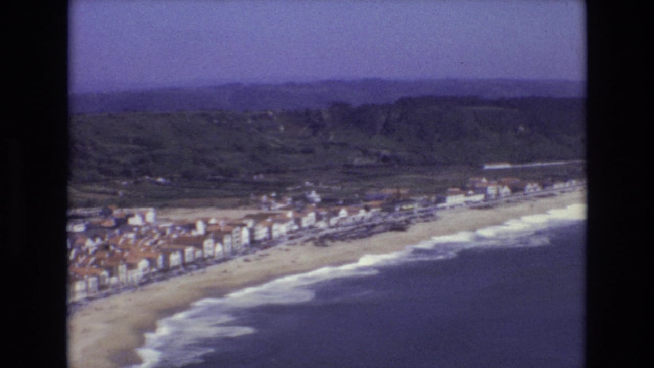 1972 Portugal aerial frame showing the coastline from above, with beachfront buildings, a wide beach, rolling surf, and inland green hills.