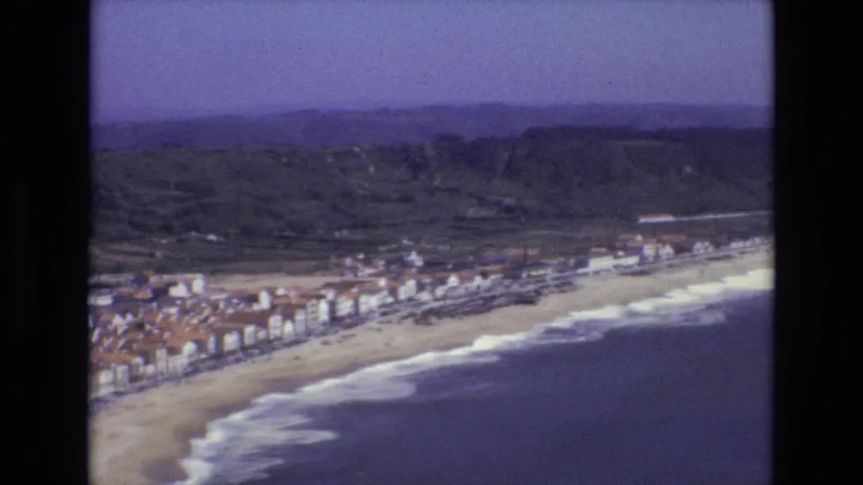 1972 Portugal aerial frame showing the coastline from above, with beachfront buildings, a wide beach, rolling surf, and inland green hills.