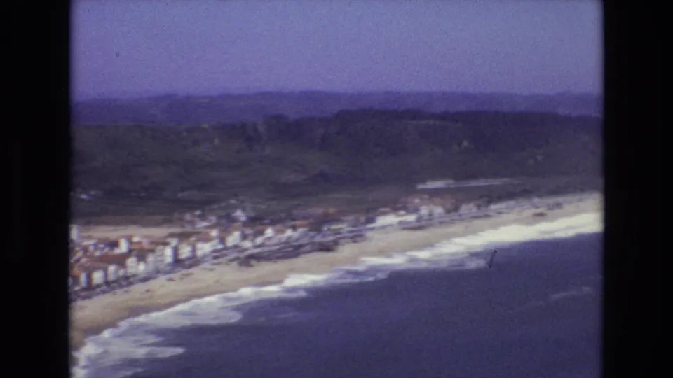 Soft 8mm aerial frame from Portugal in 1972, looking over a long sandy beach with surf, seafront structures, and hills under a purple-blue sky.