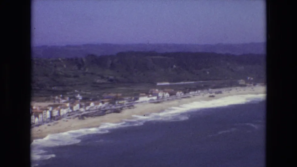1972 Portugal 8mm aerial view looking down the coast, with blue sea, white surf, sandy beach, town buildings, and green hills in the distance.