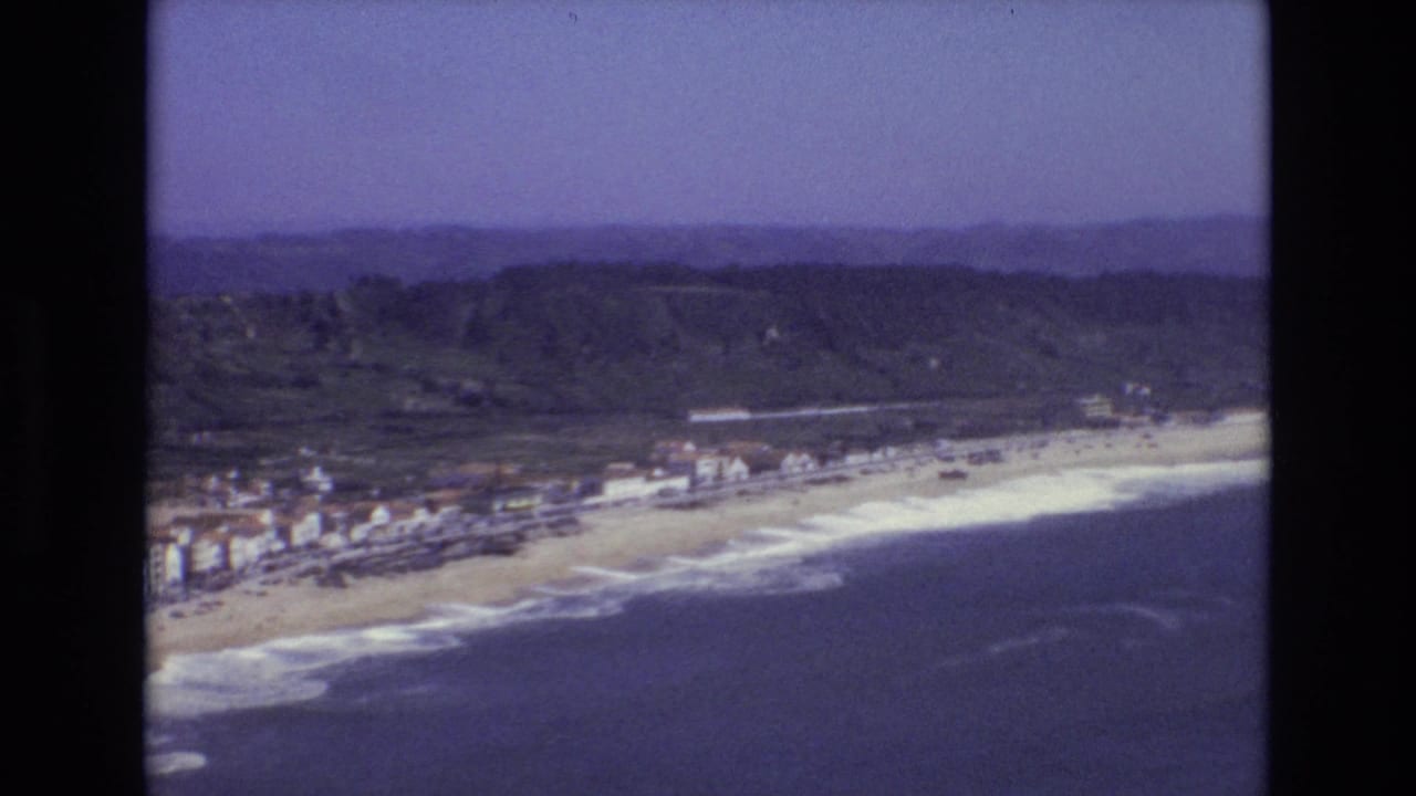 Aerial view of a sandy coastline with ocean, surf, buildings, hills, and blue sky.
