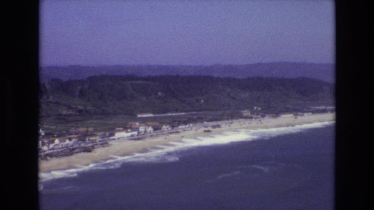 Aerial coastal view with a beach, waves, hillside, shoreline buildings, and open sky.