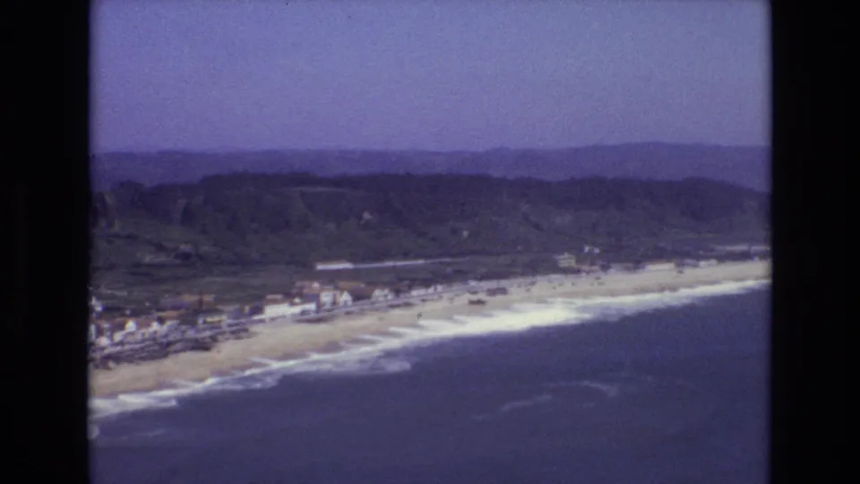 A 1972 Portugal aerial frame shows surf meeting a sandy beach below a line of coastal buildings, with wooded hills and a hazy horizon behind them.