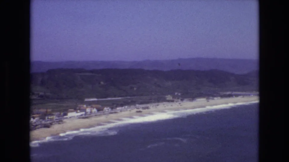 Vintage 8mm frame from Portugal in 1972 showing a long coastal beach, rolling surf, small buildings near the shore, and green hills under a pale blue sky.