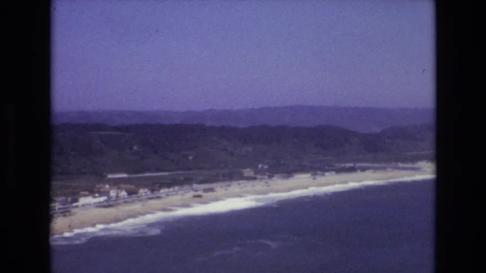 The final 1972 Portugal frame holds on the beach and ocean from above, with white surf, a coastal settlement, layered hills, and visible 8mm color fading.
