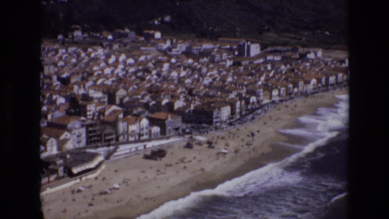 1972 Portugal 8mm frame showing an elevated view over a sandy beach, breaking waves, a waterfront promenade, and dense seaside buildings backed by hills.