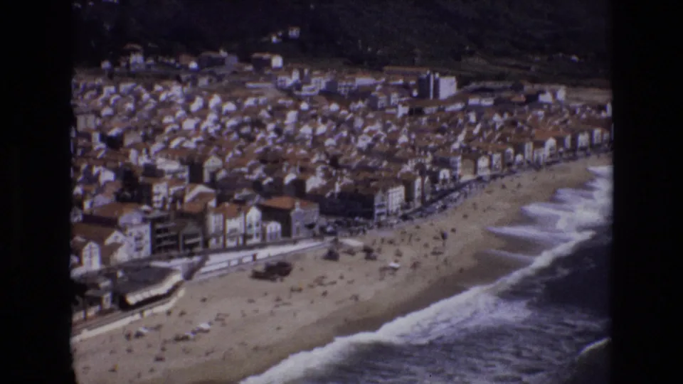 1972 Portugal 8mm frame showing an elevated view over a sandy beach, breaking waves, a waterfront promenade, and dense seaside buildings backed by hills.