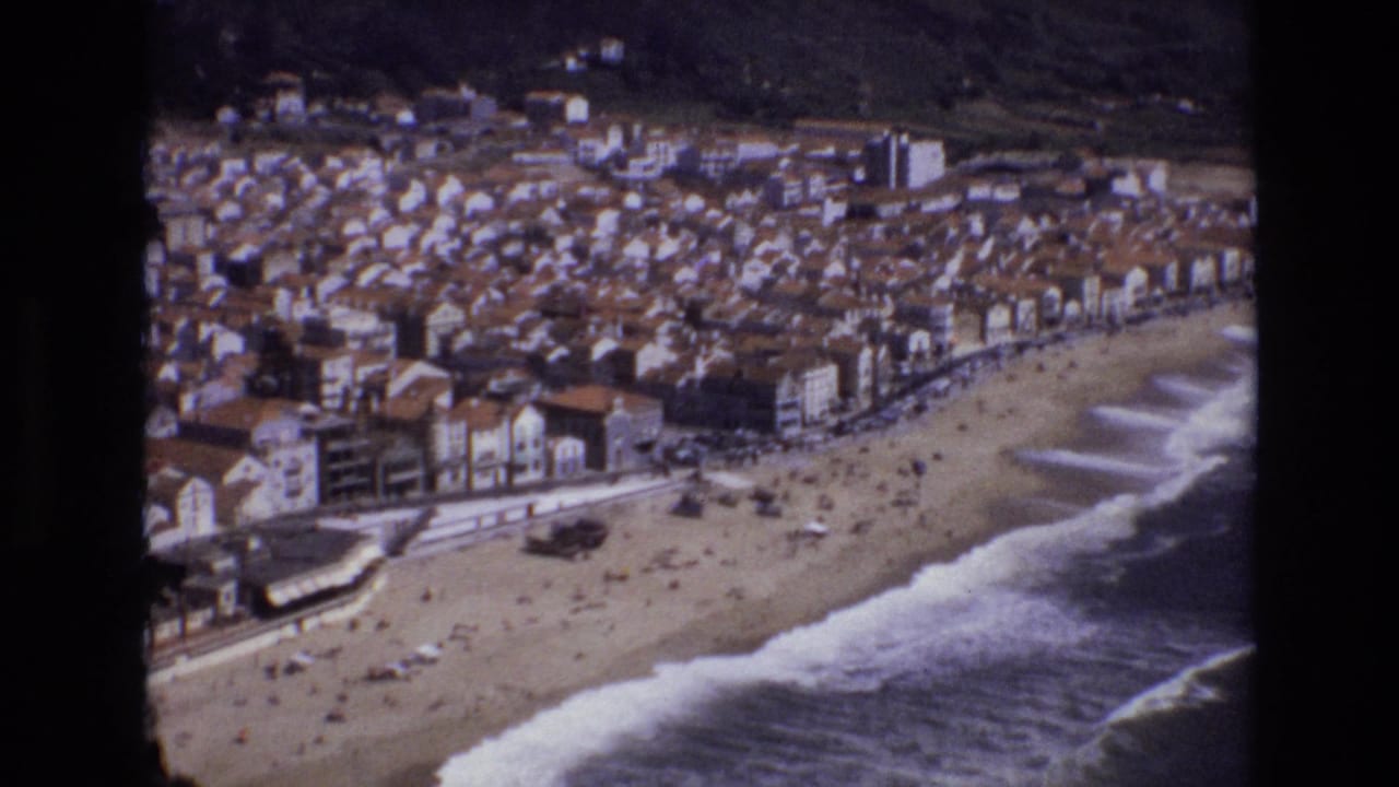 Coastal beach town seen from above with surf rolling onto the sand and people gathered along the shoreline.