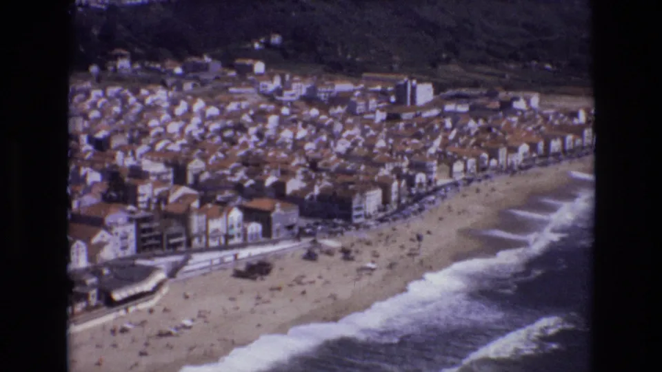 Portugal 1972 8mm still showing a tilted elevated view of a popular beach, foamy waves, a curved promenade, and compact seaside housing.