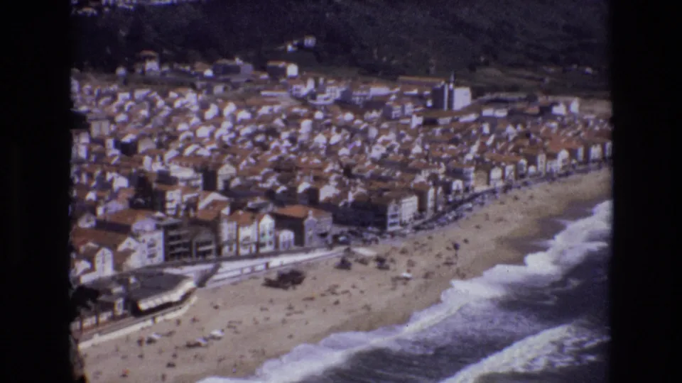 1972 Portugal vintage 8mm frame showing a broad coastal resort view with surf breaking onto sand, beachgoers, waterfront buildings, and hills behind the town.