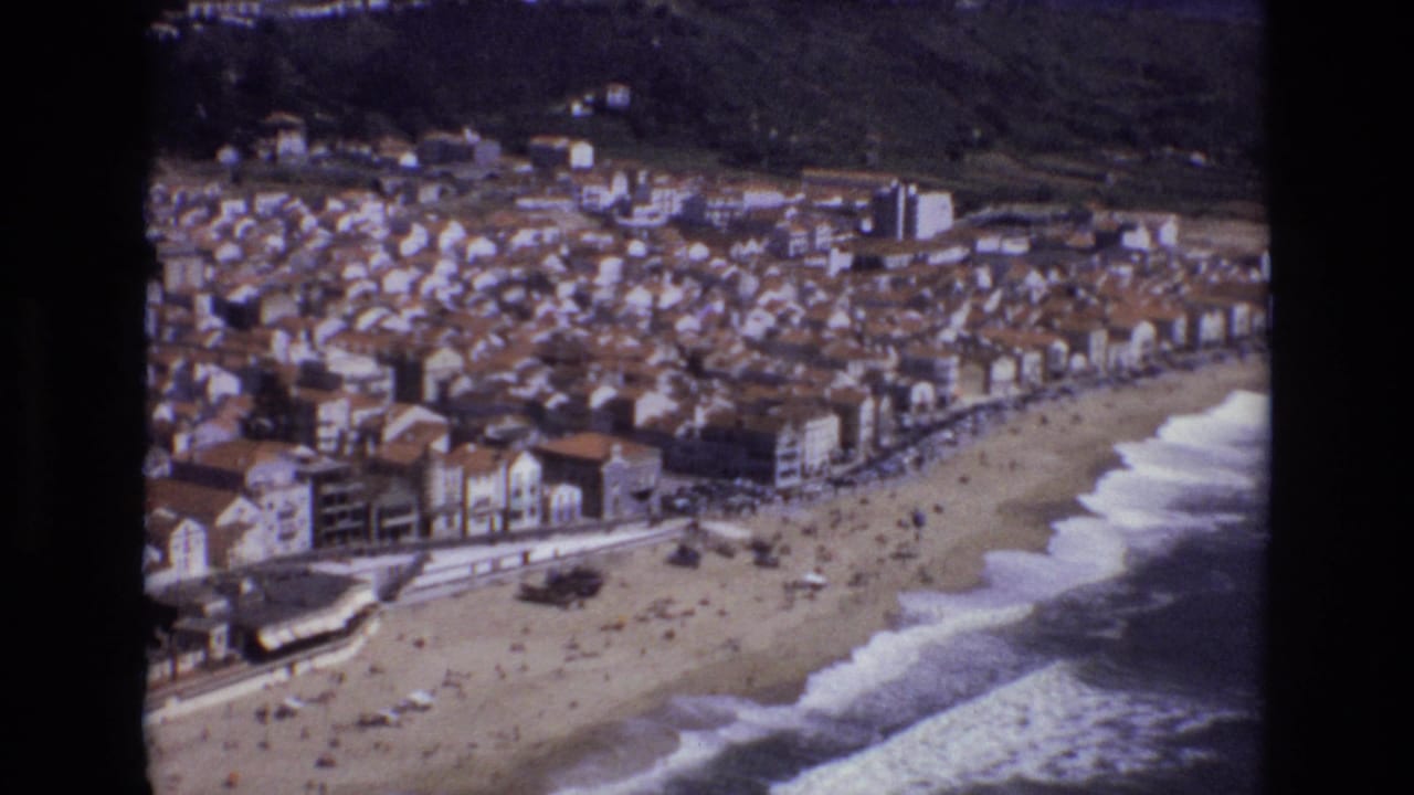 Aerial beach scene with clustered buildings, a curving shoreline, and white surf.