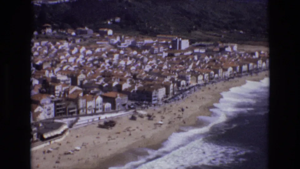 1972 Portugal 8mm aerial frame with a compact coastal settlement, terracotta-roofed buildings, a beach road, and people spread across the sand.