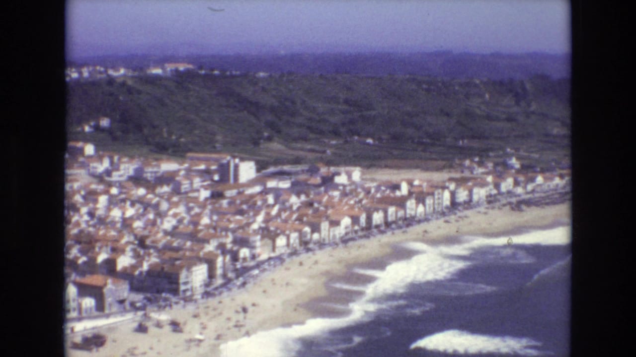 1972 Portugal 8mm aerial view of a coastal town, beachfront buildings, sandy shore, surf, green hills, and small beachgoers.