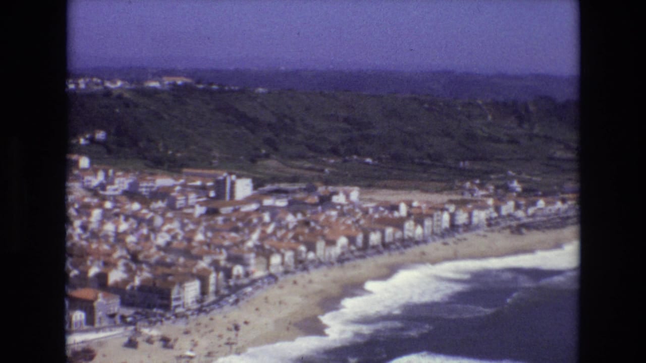 Aerial 8mm frame from Portugal in 1972 showing beachfront buildings, a sandy beach, white surf, and wooded hills beyond the town.
