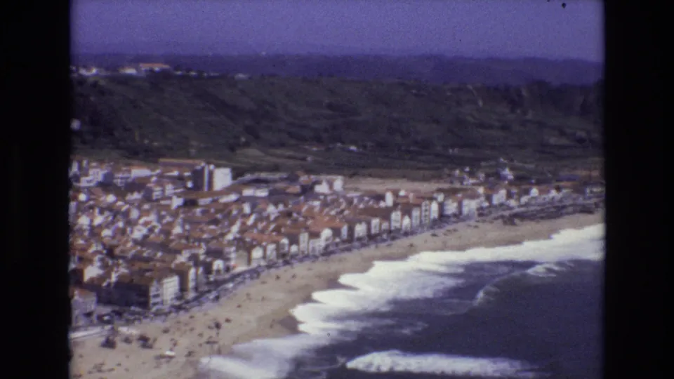 1972 Portugal aerial home-movie frame with compact coastal buildings, a broad sandy beach, breaking waves, hillside terrain, and scattered visitors.