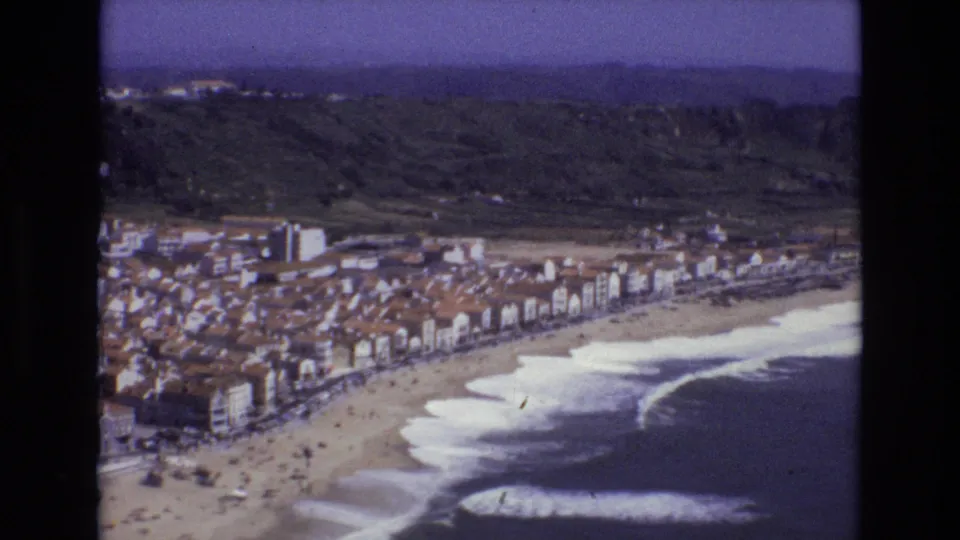 Portugal 1972 8mm aerial view of waves meeting a sandy beach below a coastal town with red roofs and green hills behind it.