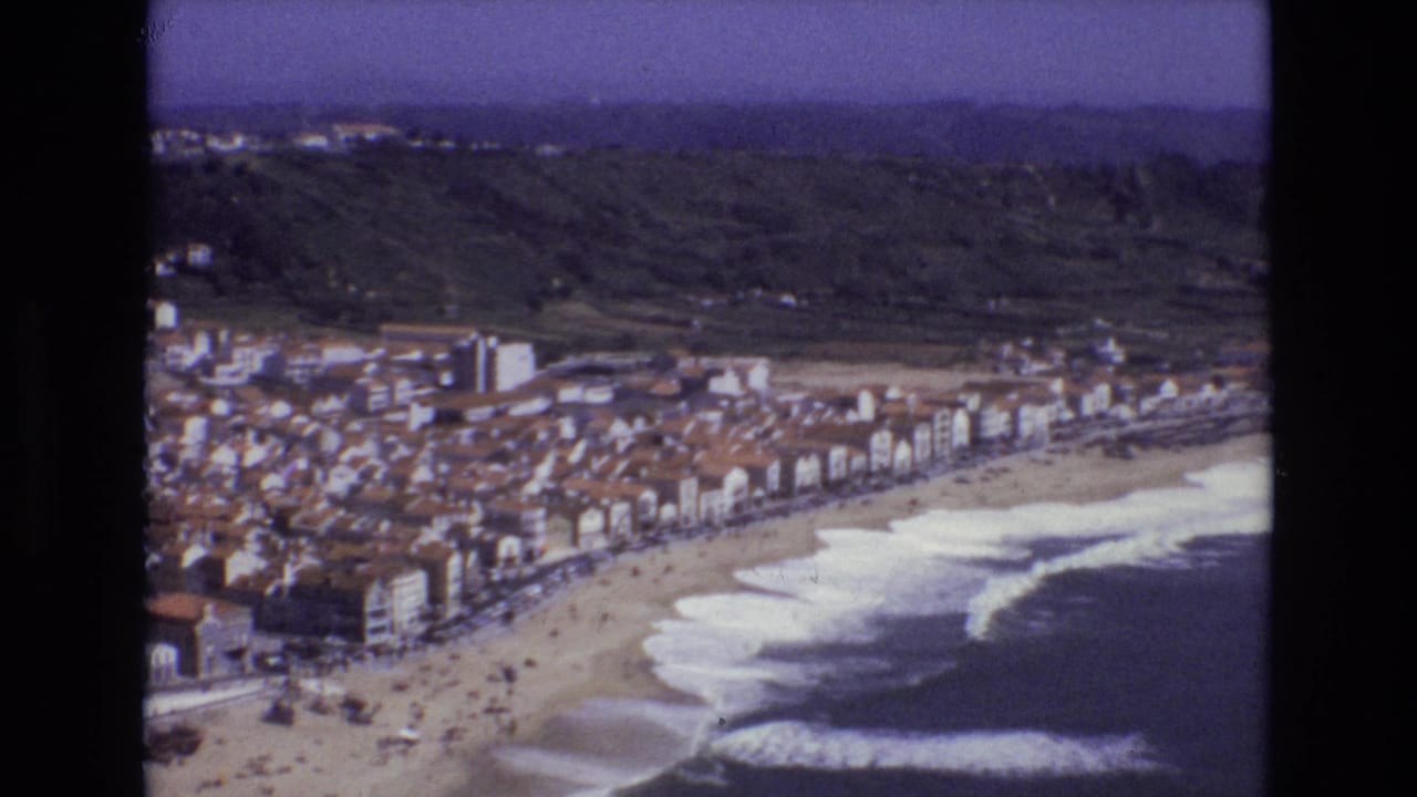 1972 Portugal 8mm frame showing a high-angle view over terracotta rooftops, beachfront buildings, a sandy shore, breaking waves, and green foothills.