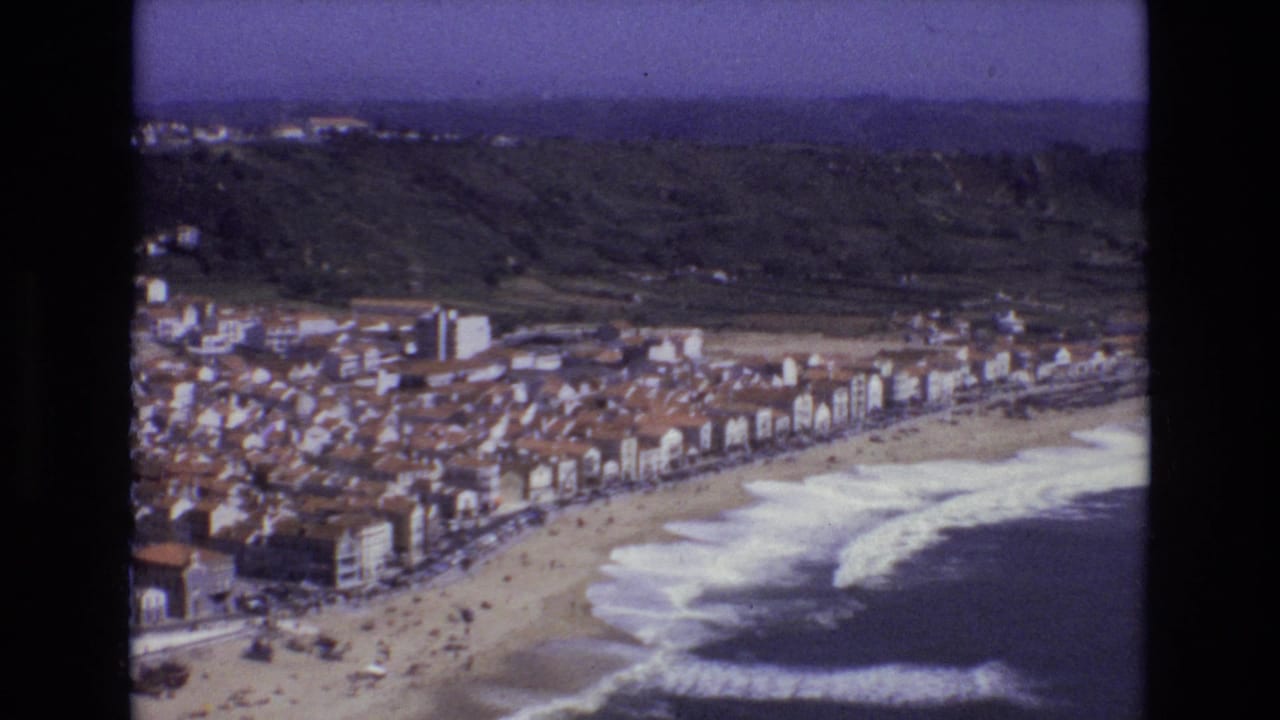 1972 Portugal 8mm frame of a seaside town lining a broad beach, with white surf rolling in and dark green foothills beyond the rooftops.