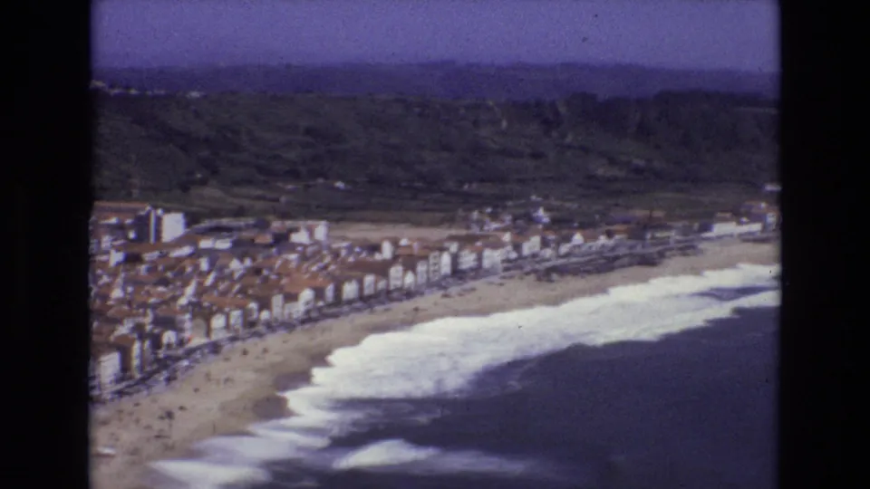1972 Portugal 8mm frame looking across a packed seaside town, a pale sand beach, foamy surf, and sloping green terrain inland.
