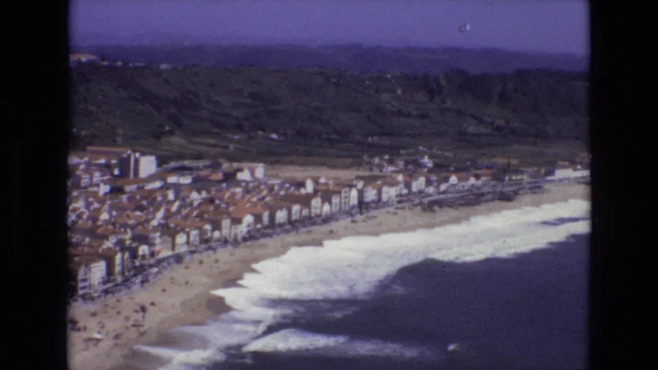 1972 Portugal 8mm frame showing a coastal town between foaming surf and green foothills, with small people visible on the beach below.