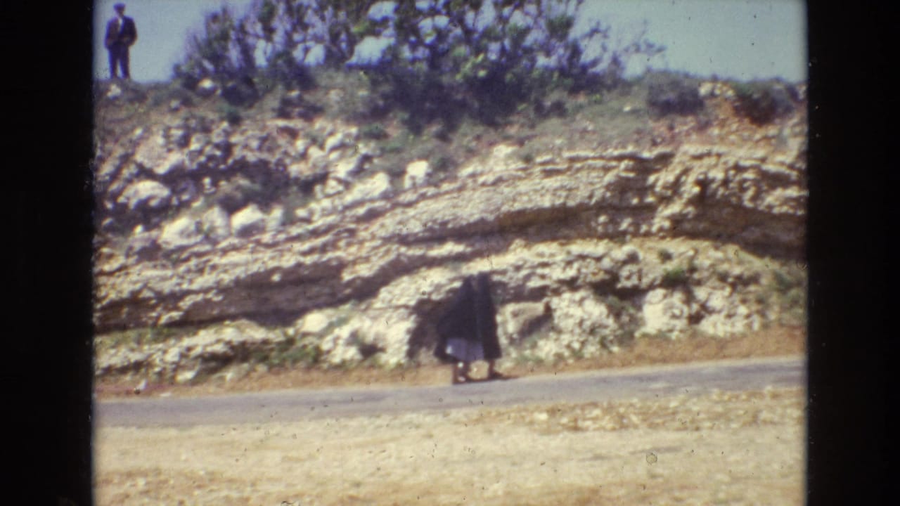 Woman in dark clothing walking along a road below a layered rocky slope.