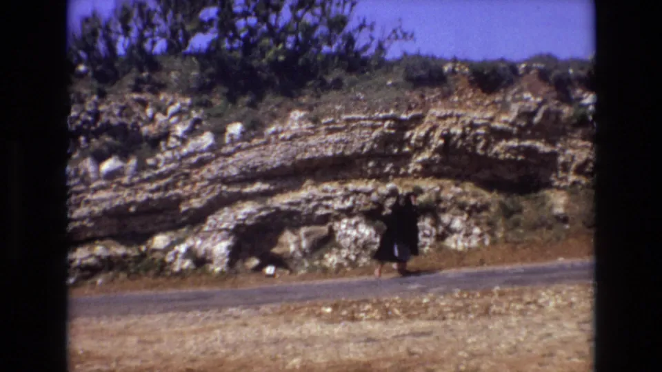 A woman in dark clothing moves along the road edge, with pale dry ground in front and a rugged layered rock slope behind her.