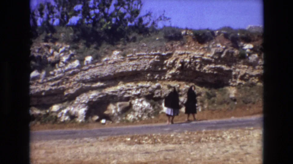 Two women in dark dresses and head coverings walk side by side on a quiet rural road in Portugal, framed against dry ground, rock strata, and sparse shrubs.
