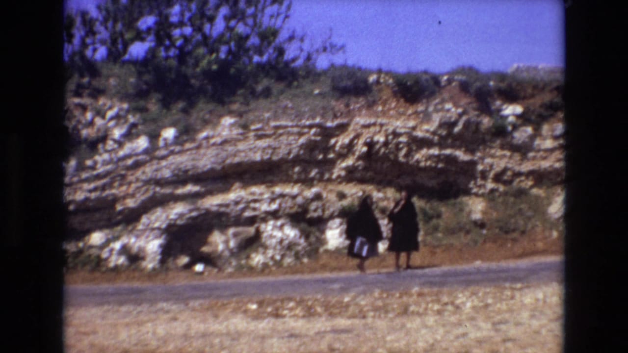In Portugal 1972 home-movie footage, two women in black cloak-like garments walk along a roadside beneath pale layered rock and sparse greenery.