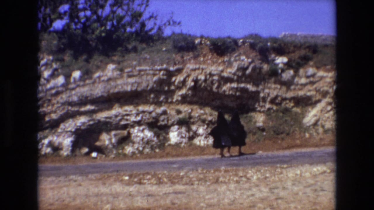 The two women remain side by side on the rural road, framed at a distance against pale rock strata and low vegetation in the 1972 Portugal footage.
