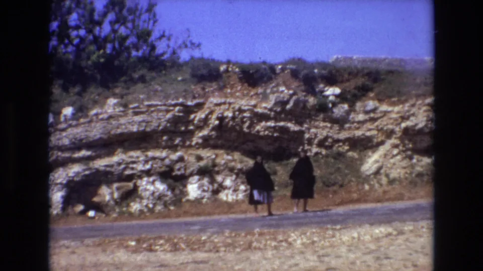 Two women in black cloak-like outerwear are seen near the road, with the rocky hillside and sparse shrubs filling the background of the 8mm Portugal scene.