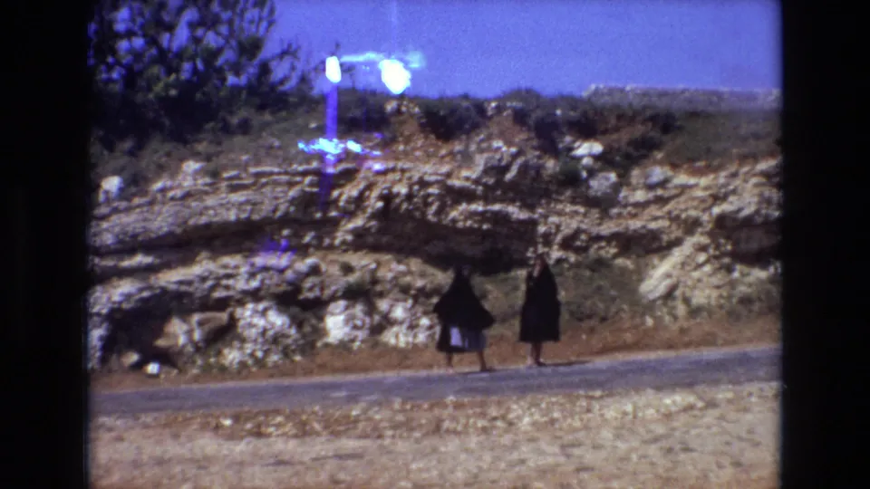 At the end of the clip, two women in dark cloak-like garments move along the roadside beneath layered rocks, with a bright film artifact visible near the upper frame.