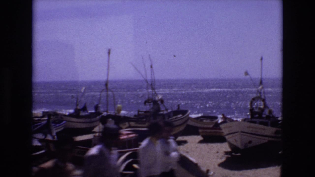 1972 Portugal home-movie frame showing small wooden boats pulled onto a sandy shore, masts and rigging against the sea, and blurred pedestrians crossing the foreground.