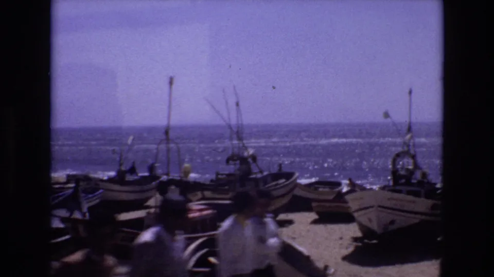 1972 Portugal home-movie frame showing small wooden boats pulled onto a sandy shore, masts and rigging against the sea, and blurred pedestrians crossing the foreground.