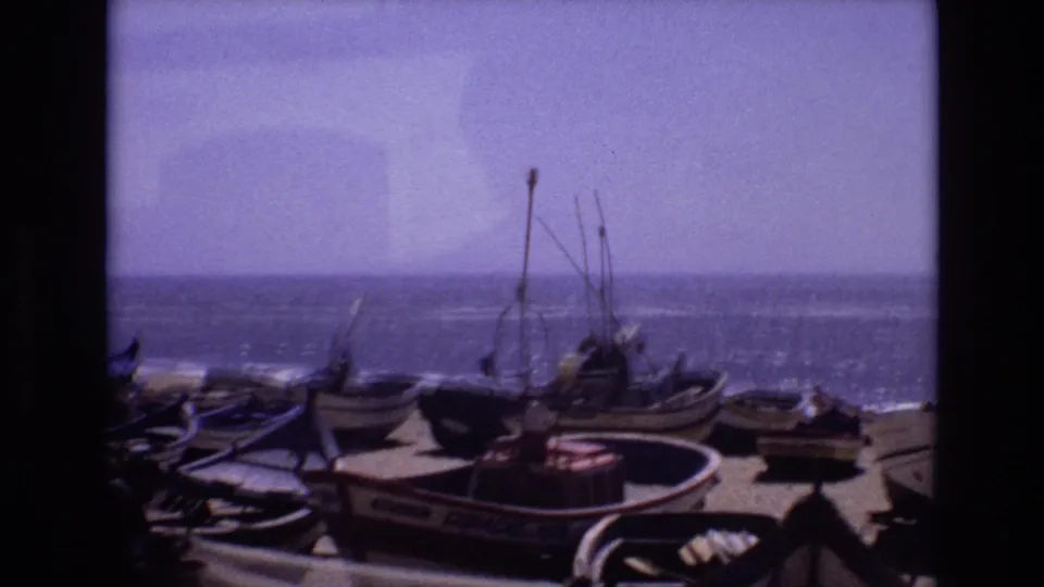 1972 Portugal coastal frame with several small boats on sand, upright rigging, painted hull lettering, and a bright ocean horizon under a pale sky.