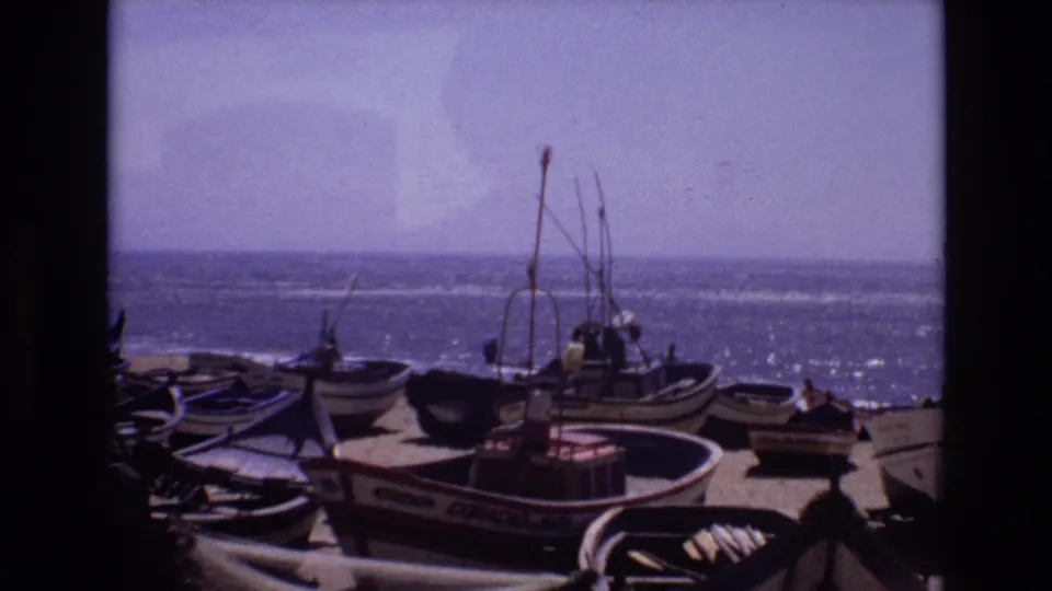 1972 Portugal home-movie frame showing a foreground boat with painted lettering, additional beached boats, upright masts, and sunlit ocean beyond the sandy shore.