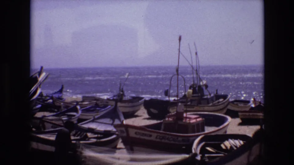 1972 Portugal coastal home-movie frame with small wooden boats on sand, visible hull lettering, masts and rigging, and the bright sea stretching across the background.