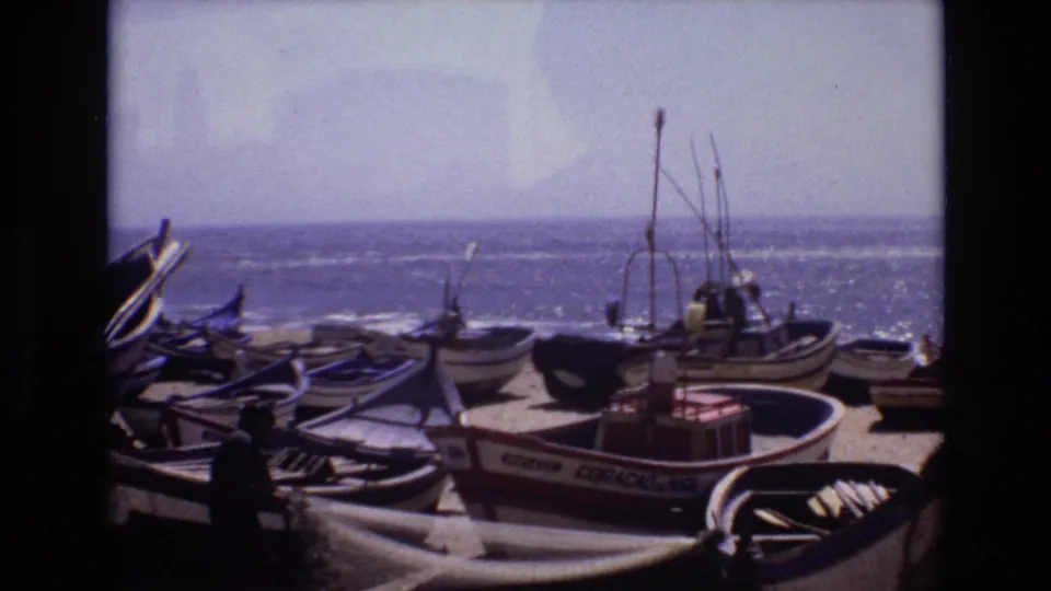 1972 Portugal 8mm frame showing small fishing boats on a sandy coastal shore, with gear in the foreground and the ocean beyond.
