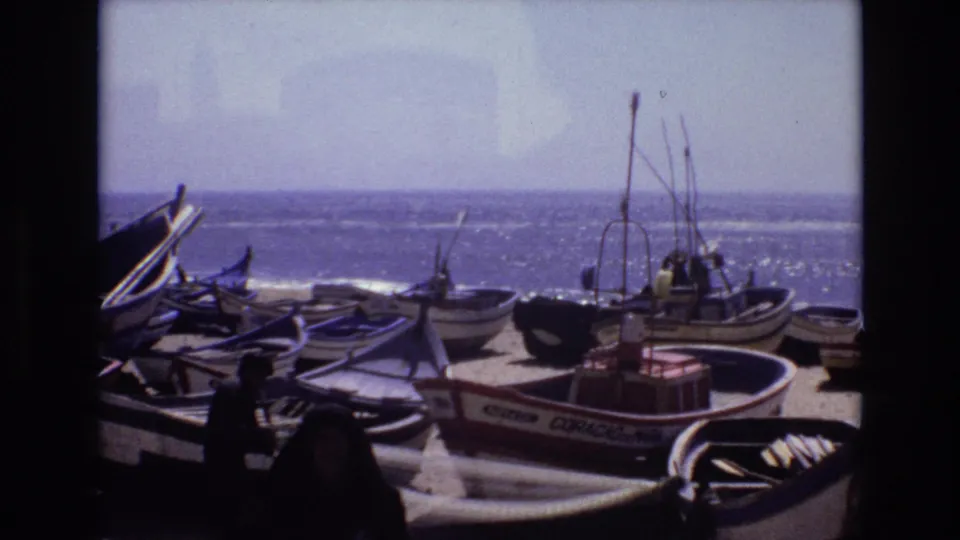 1972 Portugal 8mm frame showing small fishing boats on a sandy shore, with a dark foreground figure moving near the vessels and the ocean in the background.