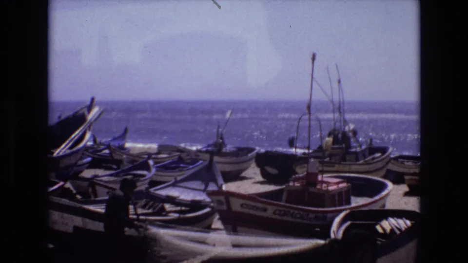 1972 Portugal home-movie frame of fishing boats arranged on sand, with a person near the lower foreground and the sea stretching behind the boats.