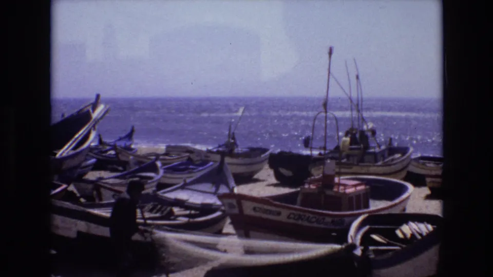 1972 Portugal home-movie frame showing wooden fishing boats pulled onto a sandy beach, with workers near the boats and the ocean behind them.