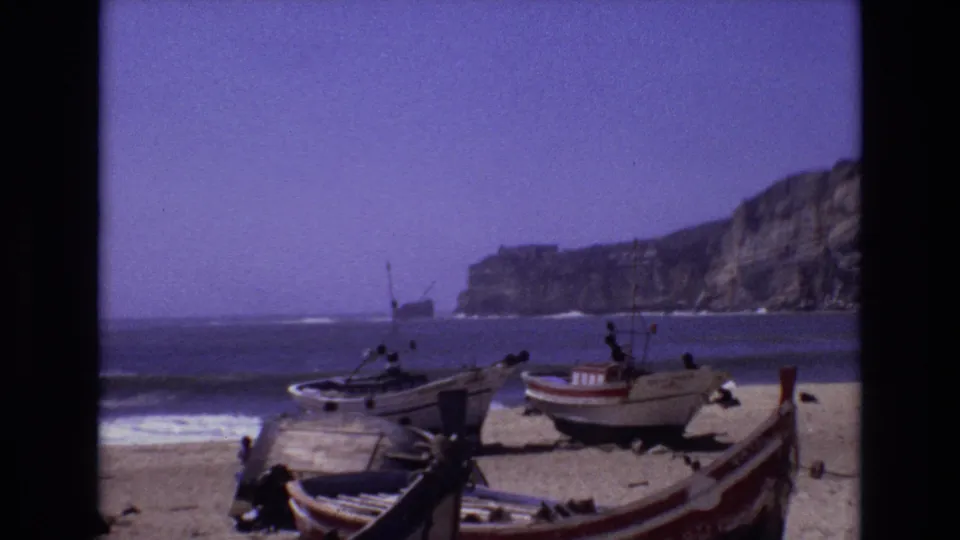 1972 Portugal 8mm frame showing several small fishing boats on a sandy beach, waves at the shoreline, and a rocky cliff rising across the bay.