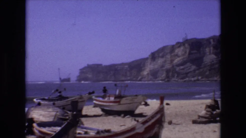 A 1972 Portugal home-movie frame shows fishing boats on the beach near low surf, with a rocky coastal cliff and open blue water beyond.